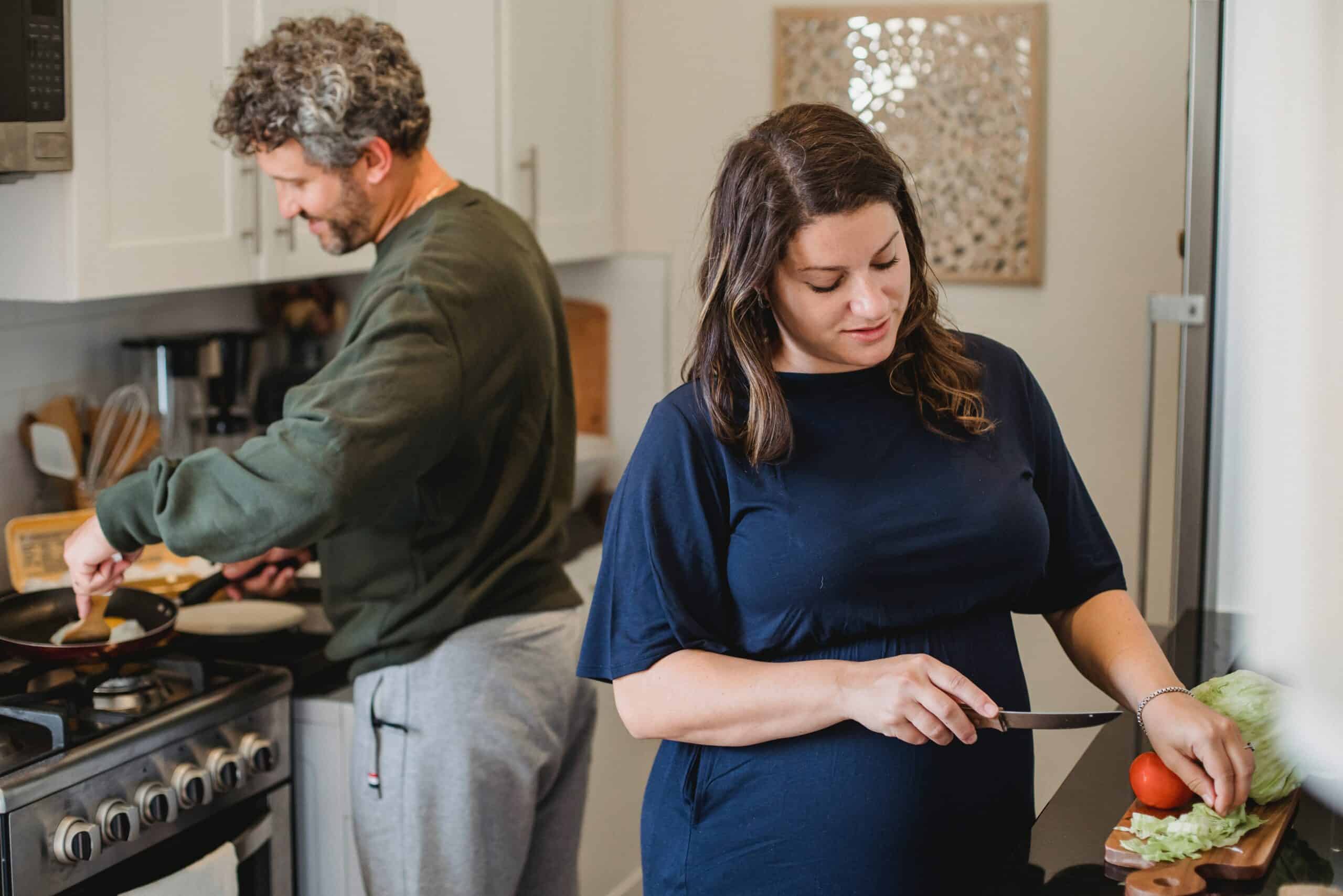 A woman with long brown hair facing cutting something on a chopping board and a man with curly here standing by the cooker and cooking