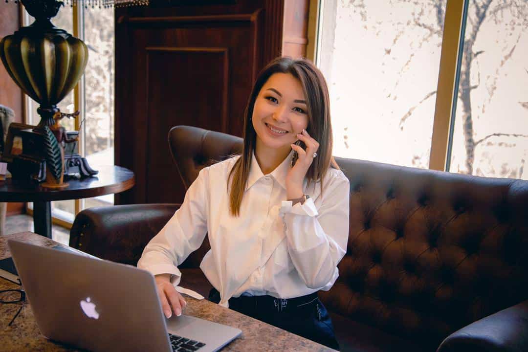 A woman with long dark hair sits at a table with a laptop in front of her, holding a mobile phone to her ear, smiling. She wears a white shirt.