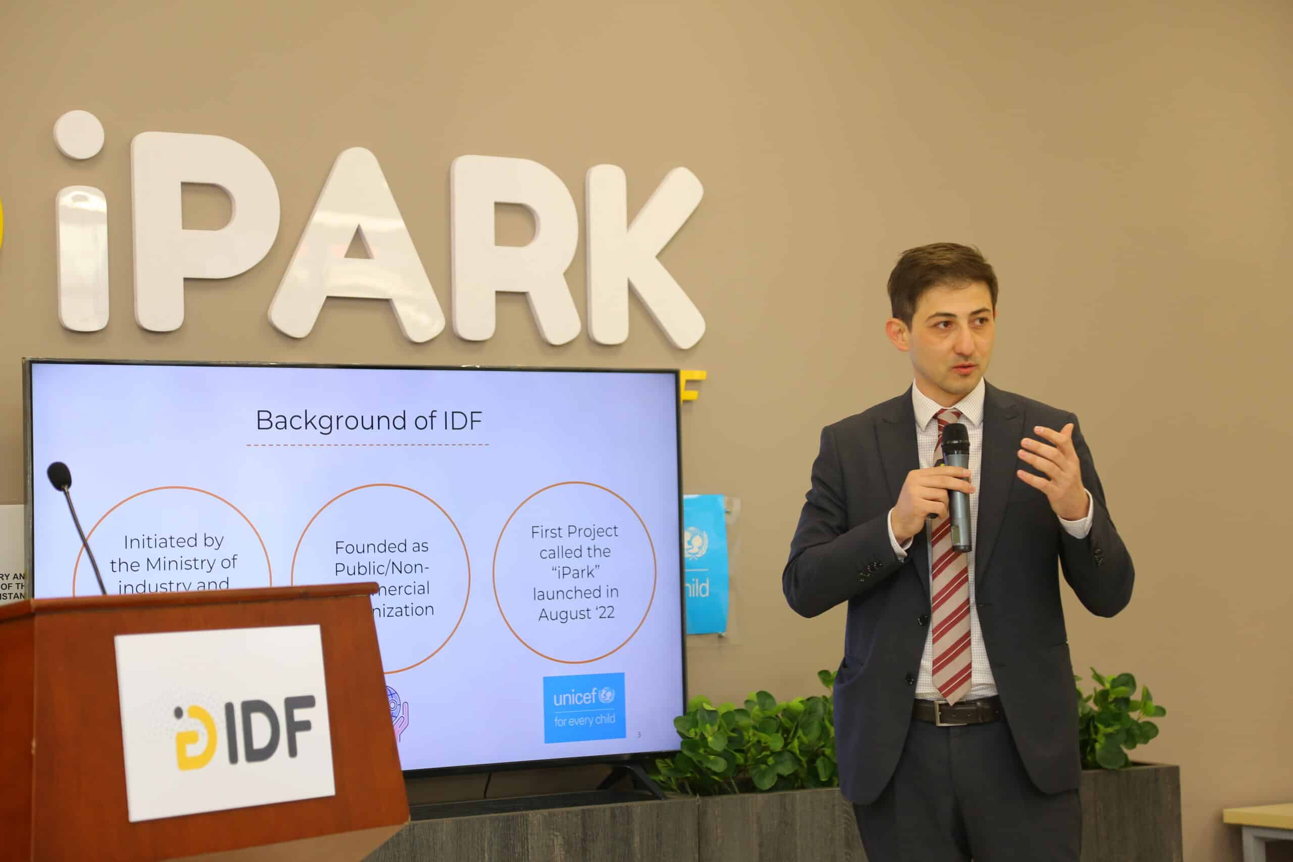 A man holding a microphone, talking, next to a screen and a sign iPark.