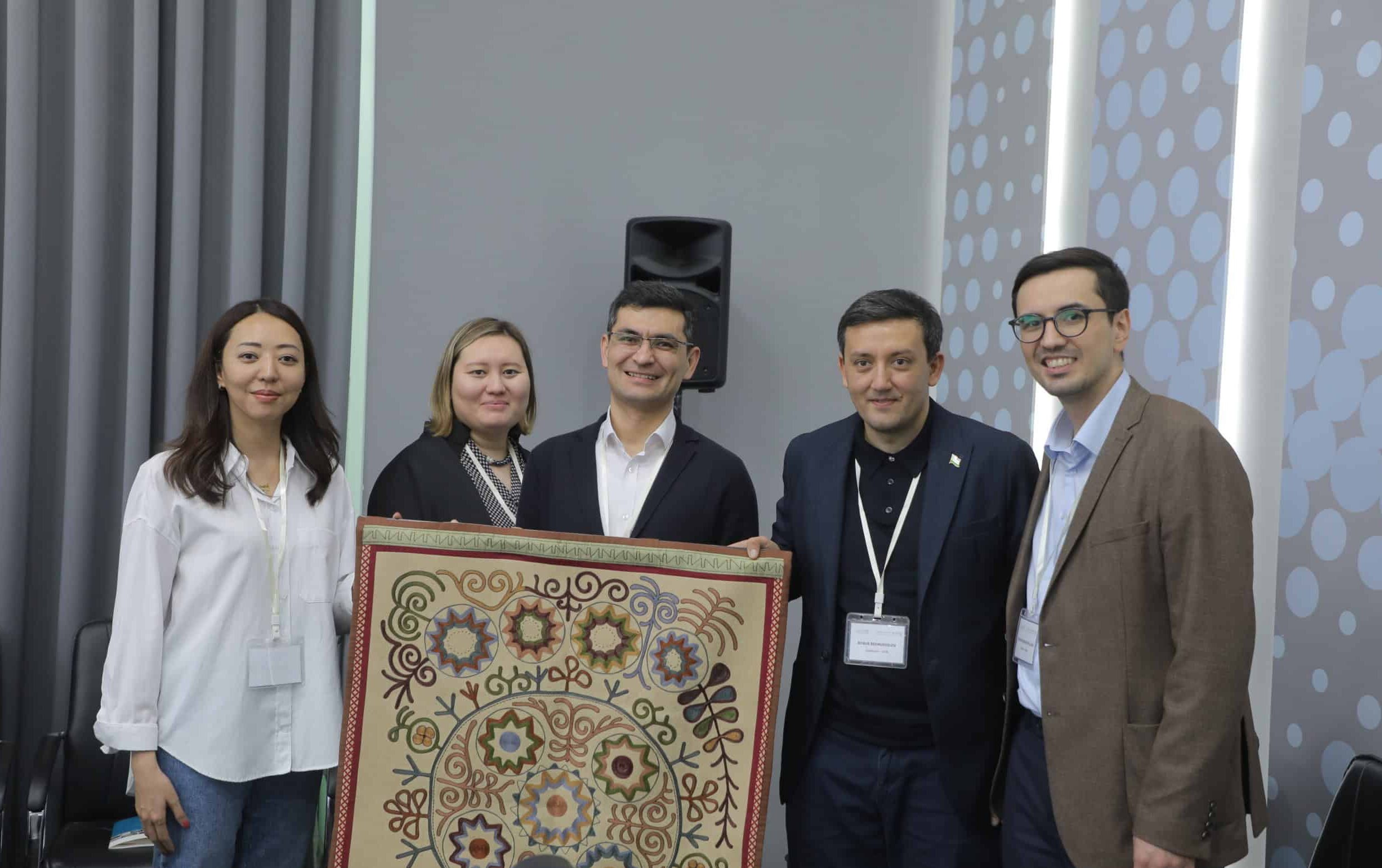 A group of people standing in a row, smiling, holding a wall hanging/rug