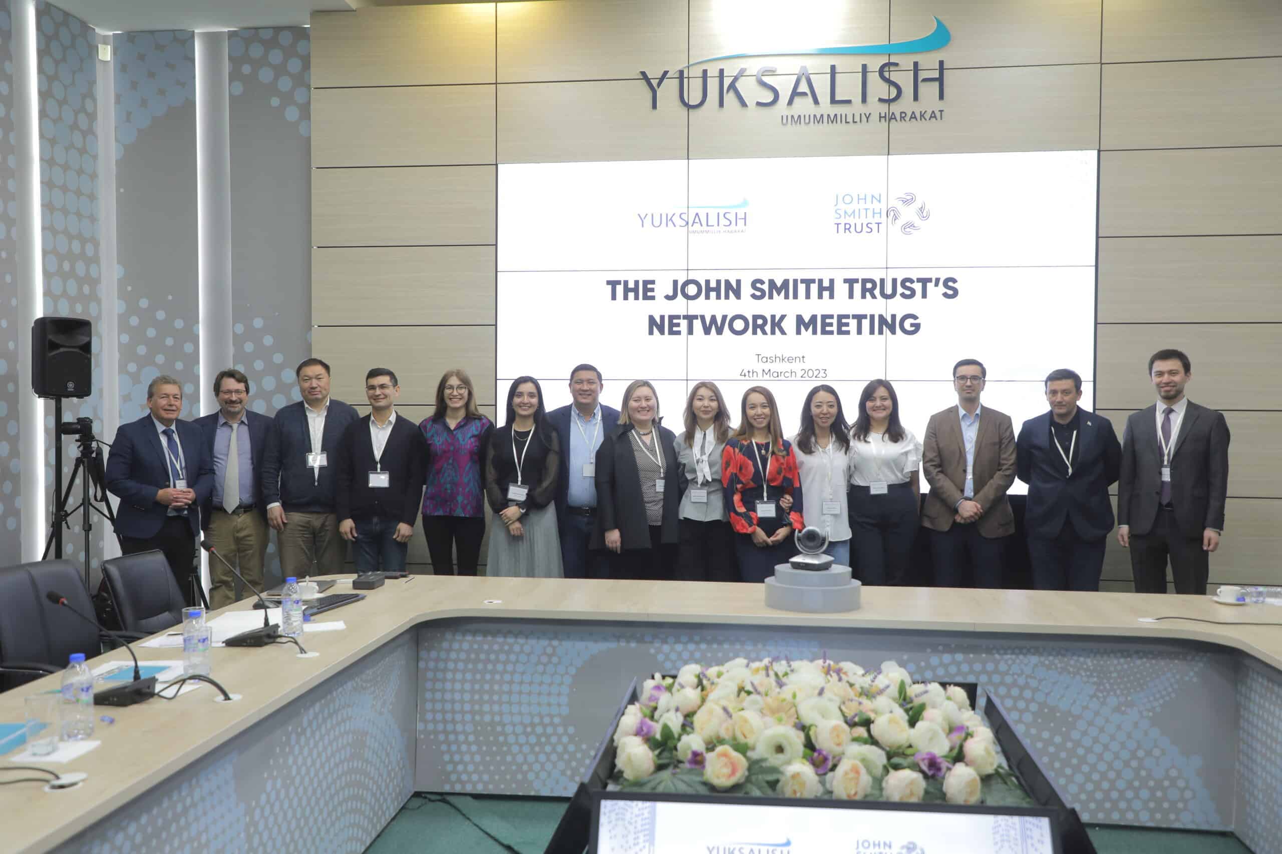 People standing at a desk with a screen behind them saying The John Smith Trust Network Meeting and Yuksalish above the screen