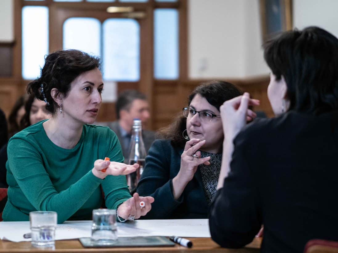 Three women at a table, one of them wearing green jumper, others wearing black. The woman in green jumper is talking and the other two are listening.