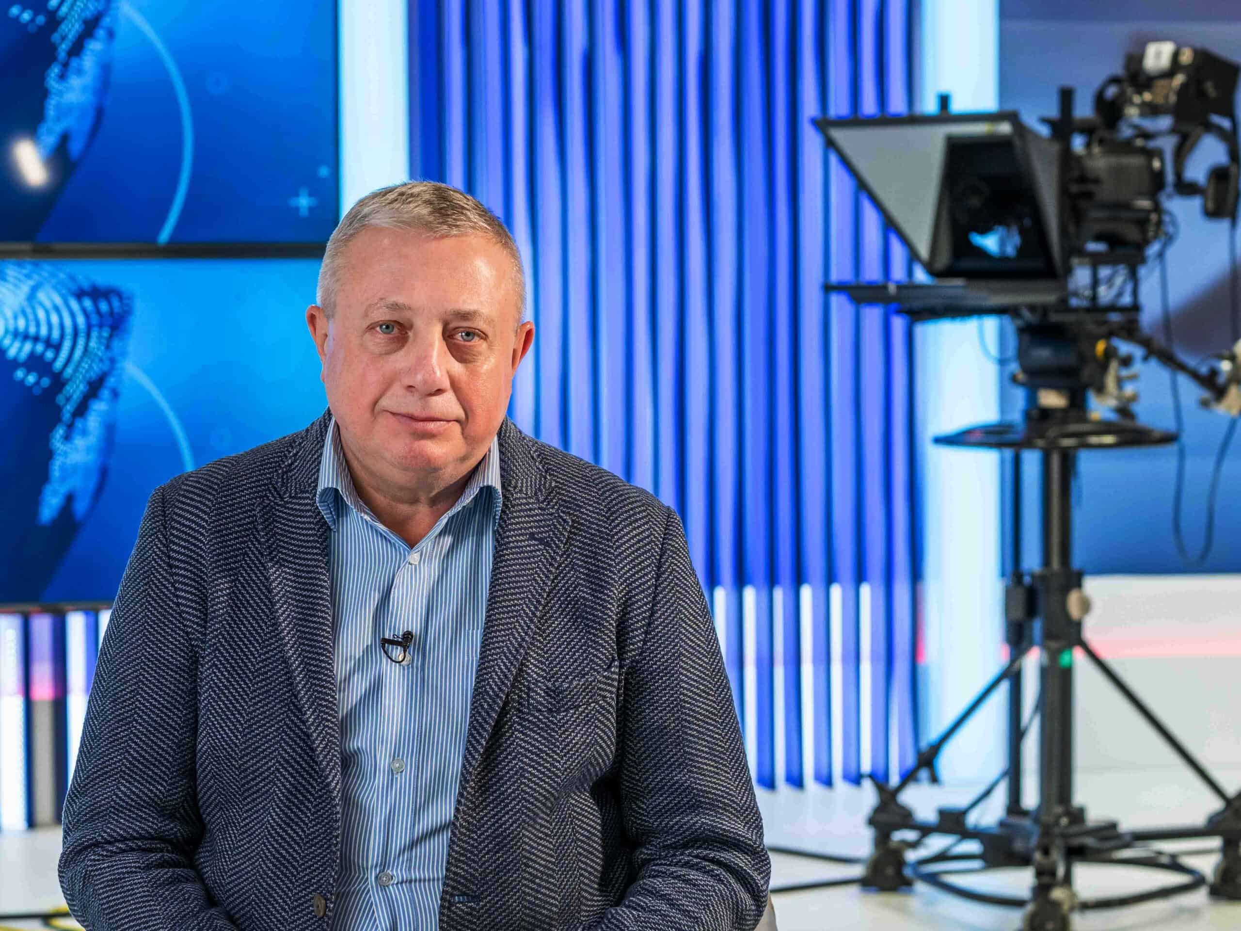 A man wearing a jacket sitting in a TV studio with a camera behind him