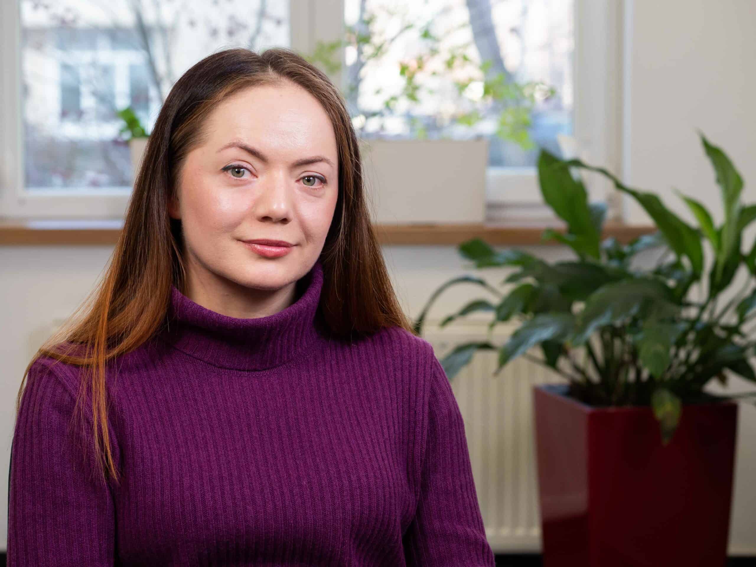 An image of a woman in purple jumper with long hair, smiling, a plant in a pot next to her and a window behind her.