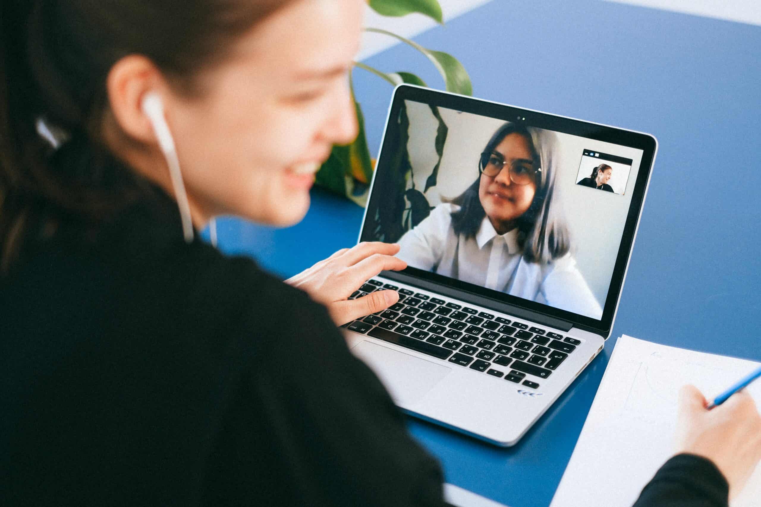 A woman sitting in front of a notebook, with a headphone in her ear, her head turned to us, another woman on the screen of the notebook smiling, they are on a video call