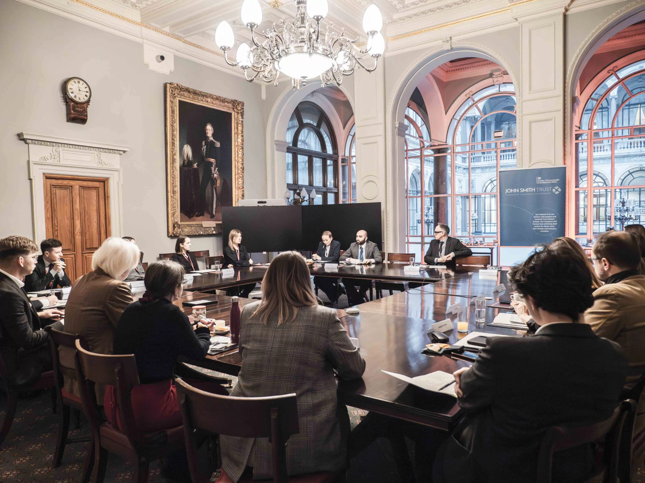 A group of people sat around a large table in a grand room with big windows and paintings